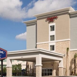 Exterior of a Hampton Inn by Hilton hotel with a covered drop-off entrance, beige and stone facade, and a blue Hampton Inn sign nearby.