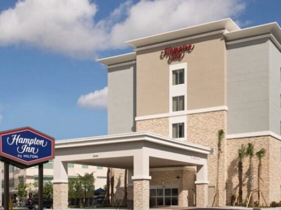 Exterior of a Hampton Inn by Hilton hotel with a covered drop-off entrance, beige and stone facade, and a blue Hampton Inn sign nearby.