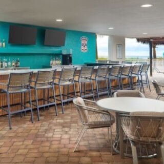 Outdoor bar area with a long high counter and metal stools, turquoise wall, bottle display, and ocean view beyond the deck.