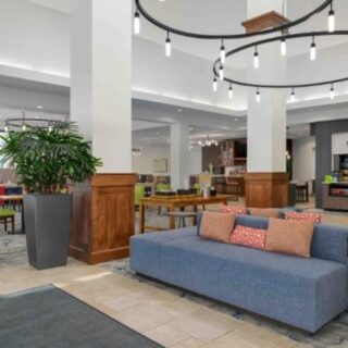 Hotel lobby lounge with a blue sofa, colorful cushions, potted plant, wood paneling, and a circular chandelier overhead.