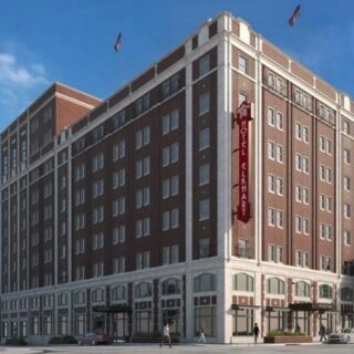 Corner view of a tall brick hotel building with a vertical red sign along the facade, blue sky above and storefronts on the ground floor.