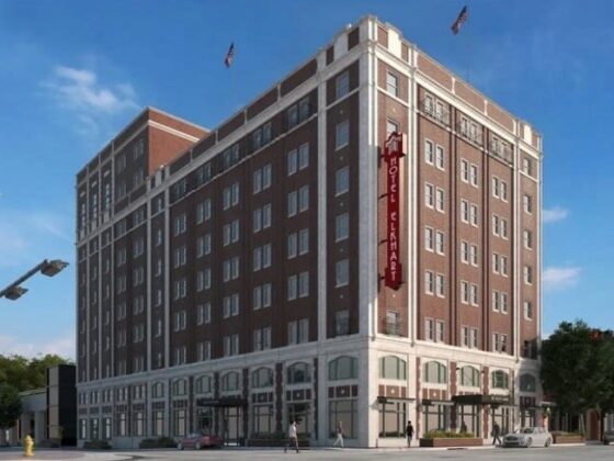 Corner view of a tall brick hotel building with a vertical red sign along the facade, blue sky above and storefronts on the ground floor.