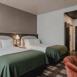 Hotel room with two double beds, white linens, and green bed runners; bedside lamps, a chair, and a doorway in view.