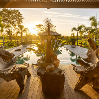 Sunset view of a seating area with carved driftwood chairs around a wooden deck, overlooking a pool and tropical garden.