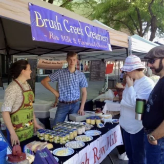 Market stall under a blue banner reading Brush Creek Creamery offering raw milk and farmstead cheese, with customers chatting outside a row of tents.