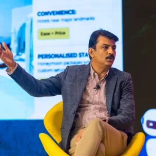 Man in a blazer sits in a bright yellow chair on stage, gesturing with his left hand during a talk, microphone clipped to shirt