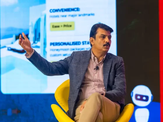 Man in a blazer sits in a bright yellow chair on stage, gesturing with his left hand during a talk, microphone clipped to shirt
