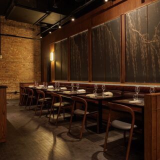 Restaurant dining area with wooden tables and chairs, a tufted bench, wine glasses set at each place, and a chalkboard wall along brick walls.