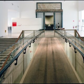 Wide indoor museum scene with a glass-railed ramp converging toward a marble doorway framed by dark columns; a person stands near the back area.
