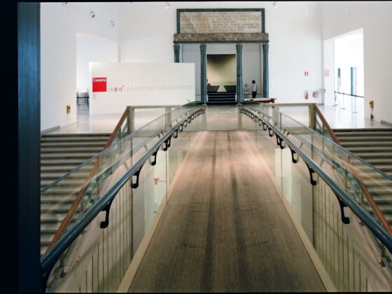 Wide indoor museum scene with a glass-railed ramp converging toward a marble doorway framed by dark columns; a person stands near the back area.
