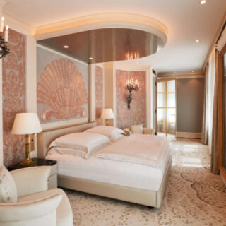 Luxurious hotel bedroom with a large upholstered bed, cream seating, and chandeliers in a gilded, patterned room.