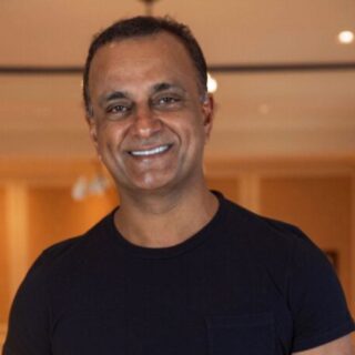 Man wearing a black t-shirt smiles at the camera in a warmly lit indoor hallway or lobby setting.