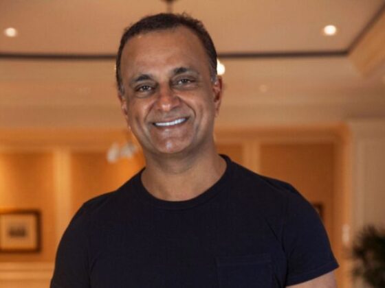 Man wearing a black t-shirt smiles at the camera in a warmly lit indoor hallway or lobby setting.