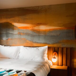 Hotel bedroom with white bedding and a desert landscape mural behind the bed, two wall lamps, and a wooden headboard.
