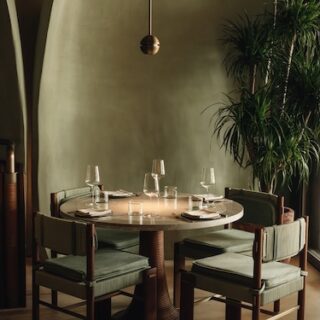 Round round dining table for four with wine glasses and napkins in a modern booth area, green upholstered chairs, and a hanging pendant light nearby.