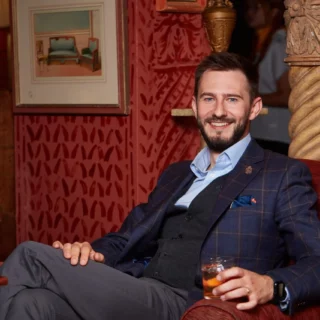 Man in a navy plaid blazer and vest sits in a red velvet chair, holding a glass and smiling at the camera.