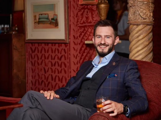 Man in a navy plaid blazer and vest sits in a red velvet chair, holding a glass and smiling at the camera.