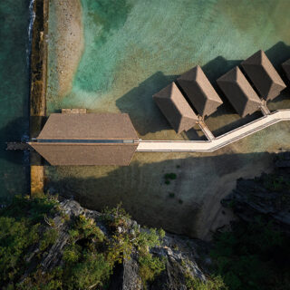 Aerial view of a thatched-roof bungalow resort with overwater villas connected by a long wooden walkway to a larger central building, along a turquoise shoreline.