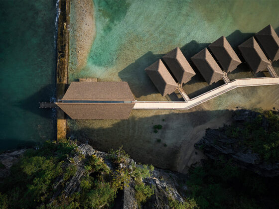 Aerial view of a thatched-roof bungalow resort with overwater villas connected by a long wooden walkway to a larger central building, along a turquoise shoreline.