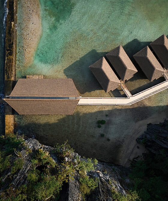 Aerial view of a thatched-roof bungalow resort with overwater villas connected by a long wooden walkway to a larger central building, along a turquoise shoreline.