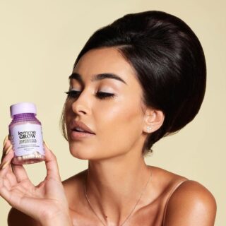Woman holding a lavender bottle labeled 'Lemme GROW' (hair growth) close to her face in a beige studio setting, with makeup and sleek dark hair.