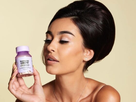 Woman holding a lavender bottle labeled 'Lemme GROW' (hair growth) close to her face in a beige studio setting, with makeup and sleek dark hair.