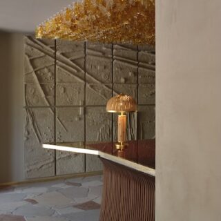 Curved wooden bar with a warm copper lamp, overhead amber glassware chandelier, and textured stone wall panels in a modern lobby.