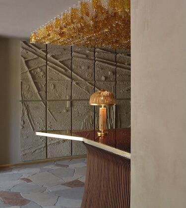 Curved wooden bar with a warm copper lamp, overhead amber glassware chandelier, and textured stone wall panels in a modern lobby.