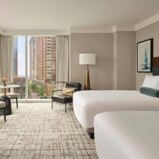 Hotel room with two double beds, seating area, and large windows overlooking a cityscape.