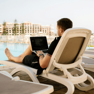 Man relaxing on a poolside lounge chair using a laptop with buildings and palm trees in the background