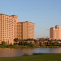 Resort hotel complex along a lake with palm trees and a grassy foreground under a clear blue sky