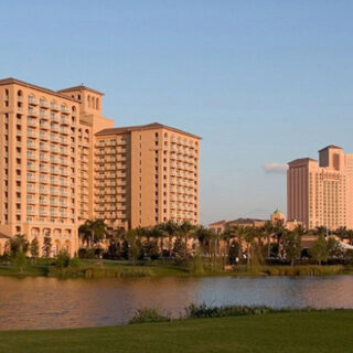 Resort hotel complex along a lake with palm trees and a grassy foreground under a clear blue sky