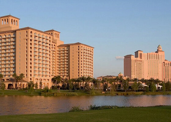 Resort hotel complex along a lake with palm trees and a grassy foreground under a clear blue sky