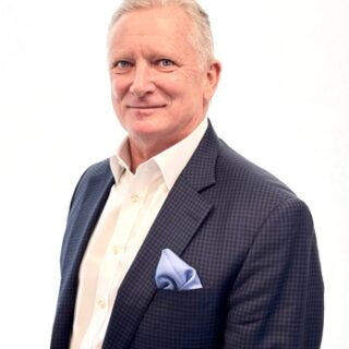Professional headshot of a mature man in a navy check blazer, white shirt, light blue pocket square, smiling confidently.