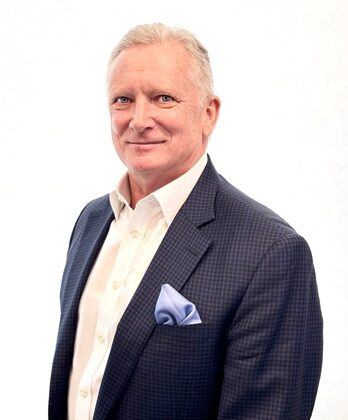 Professional headshot of a mature man in a navy check blazer, white shirt, light blue pocket square, smiling confidently.