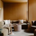 Cozy lounge area with a beige sectional, round coffee tables, and an armchair in a warm brown-walled room.",