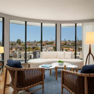 Bright living room with curved floor-to-ceiling windows overlooking a cityscape; white sofa, wicker chairs, blue rug, and wood coffee tables.