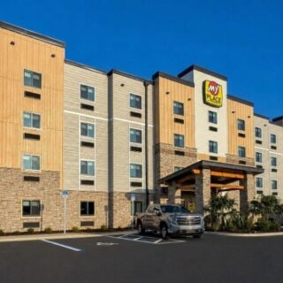 Exterior view of a My Place hotel with a beige and gray facade, palm trees, and parked cars under a clear blue sky