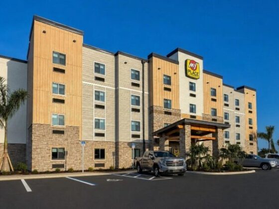 Exterior view of a My Place hotel with a beige and gray facade, palm trees, and parked cars under a clear blue sky