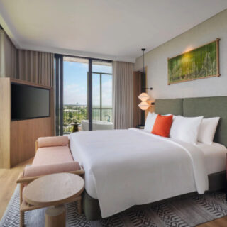 Bright hotel room with a king bed, white bedding and an orange accent pillow, green upholstered headboard, and paired nightstands with pendant lights. A flat-screen TV sits on a wooden stand opposite the bed. At the foot is a small round table and pink bench. Large windows and a glass door lead to a balcony with potted plants and a city view, creating a airy, contemporary atmosphere.
