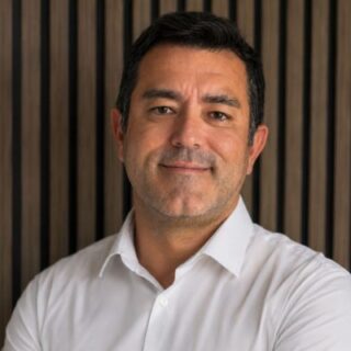 Head-and-shoulders portrait of a smiling man in a white shirt against a dark wooden panel background.