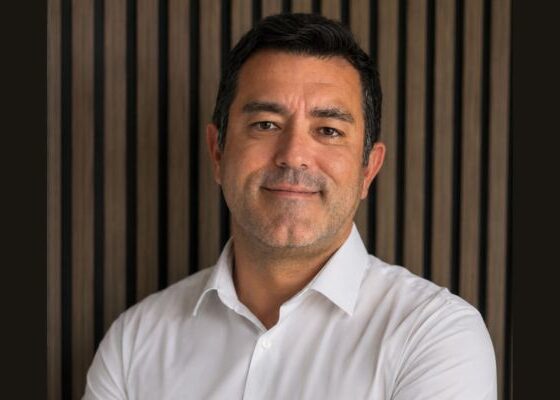 Head-and-shoulders portrait of a smiling man in a white shirt against a dark wooden panel background.