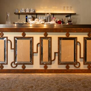 Vintage copper-framed bar counter with geometric mirrored panels and warm lighting, front view on a carpeted floor.
