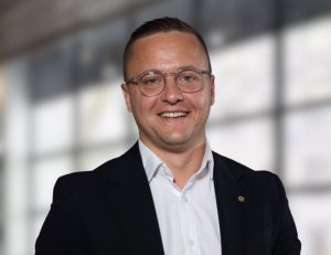 Smiling man in a navy blazer and white shirt, wearing glasses, in a bright office setting (professional headshot)