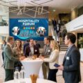 Professionals networking at round high tables in a bright modern lobby with a blue Hospitality Forum banner in the background.