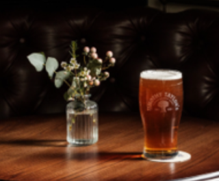 Pint of amber beer on a round wooden table with a small vase of flowers against a dark tufted background.
