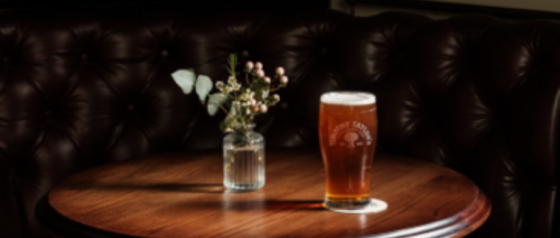 Pint of amber beer on a round wooden table with a small vase of flowers against a dark tufted background.