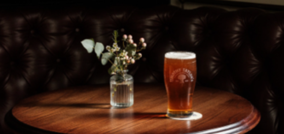Pint of amber beer on a round wooden table with a small vase of flowers against a dark tufted background.
