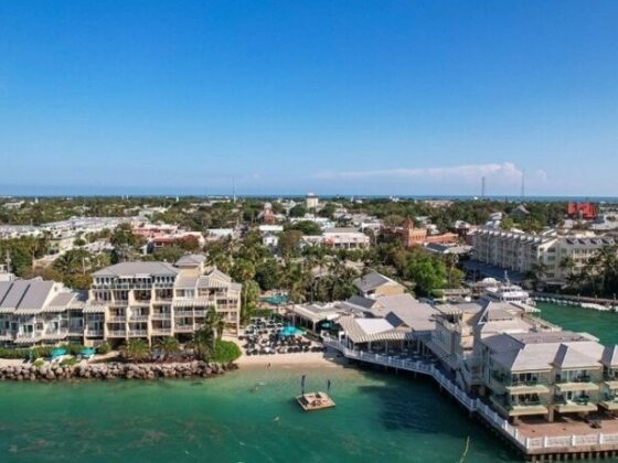 Aerial view of a tropical coastal town with waterfront resorts and a marina along clear blue-green water with palm trees.