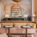 Pastel pink lounge with a curved peach sofa, floral cushions, and two round tables in front of a backlit bar.